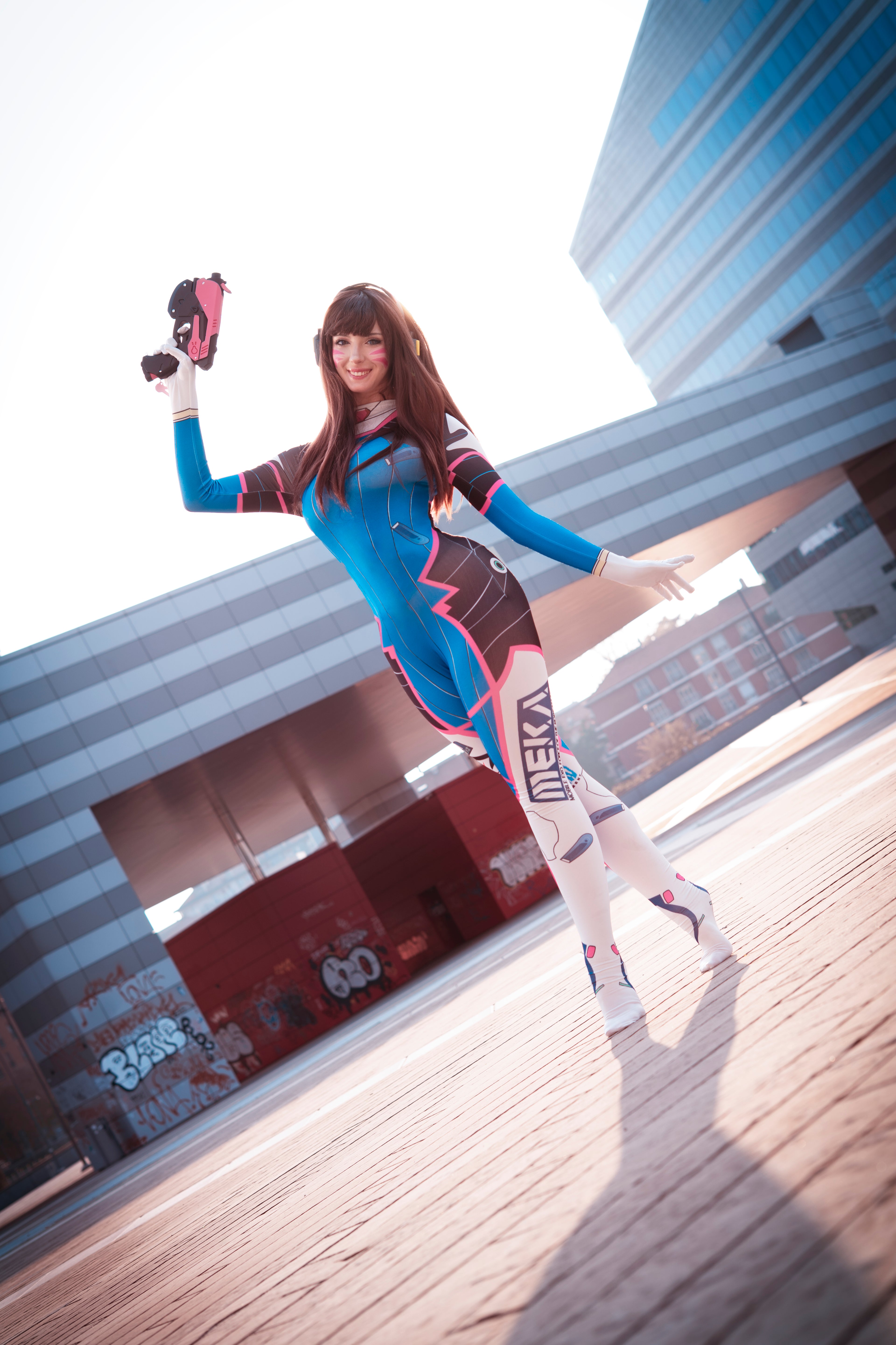 Full-length view of a cosplayer wearing the D.Va pilot suit holding a prop blaster in an urban setting